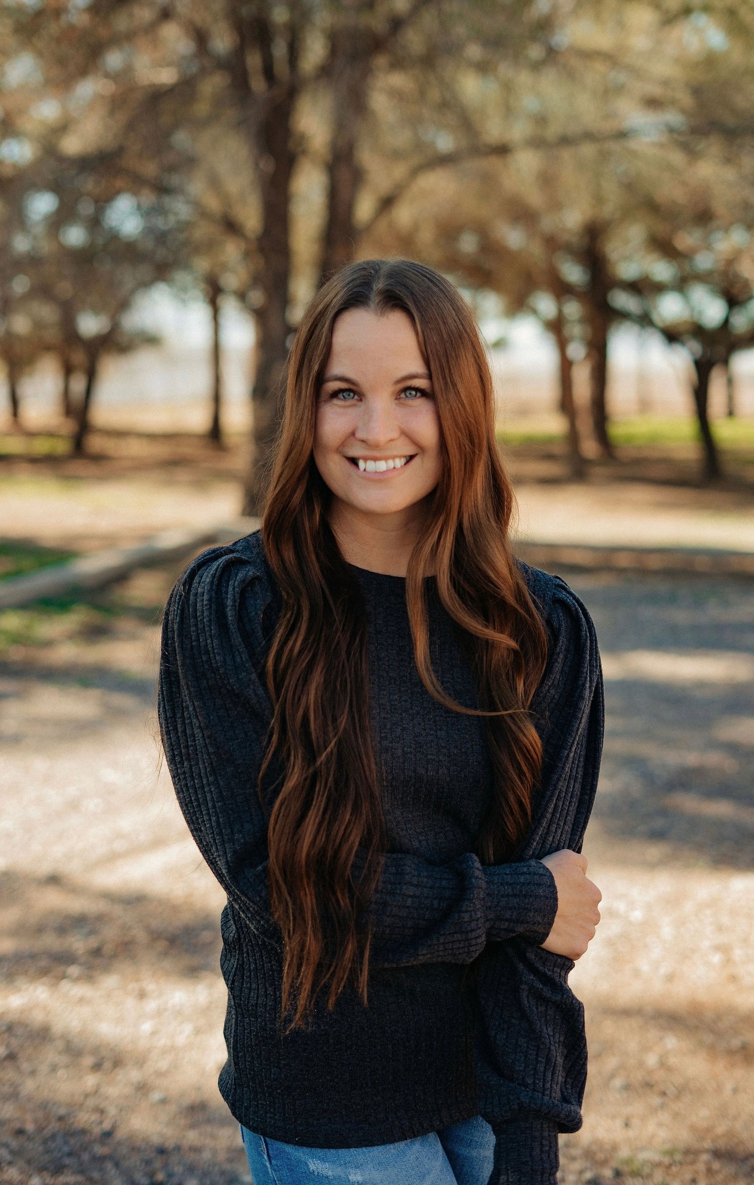 Woman with long brown hair wearing a dark sweater standing in a park-like setting.