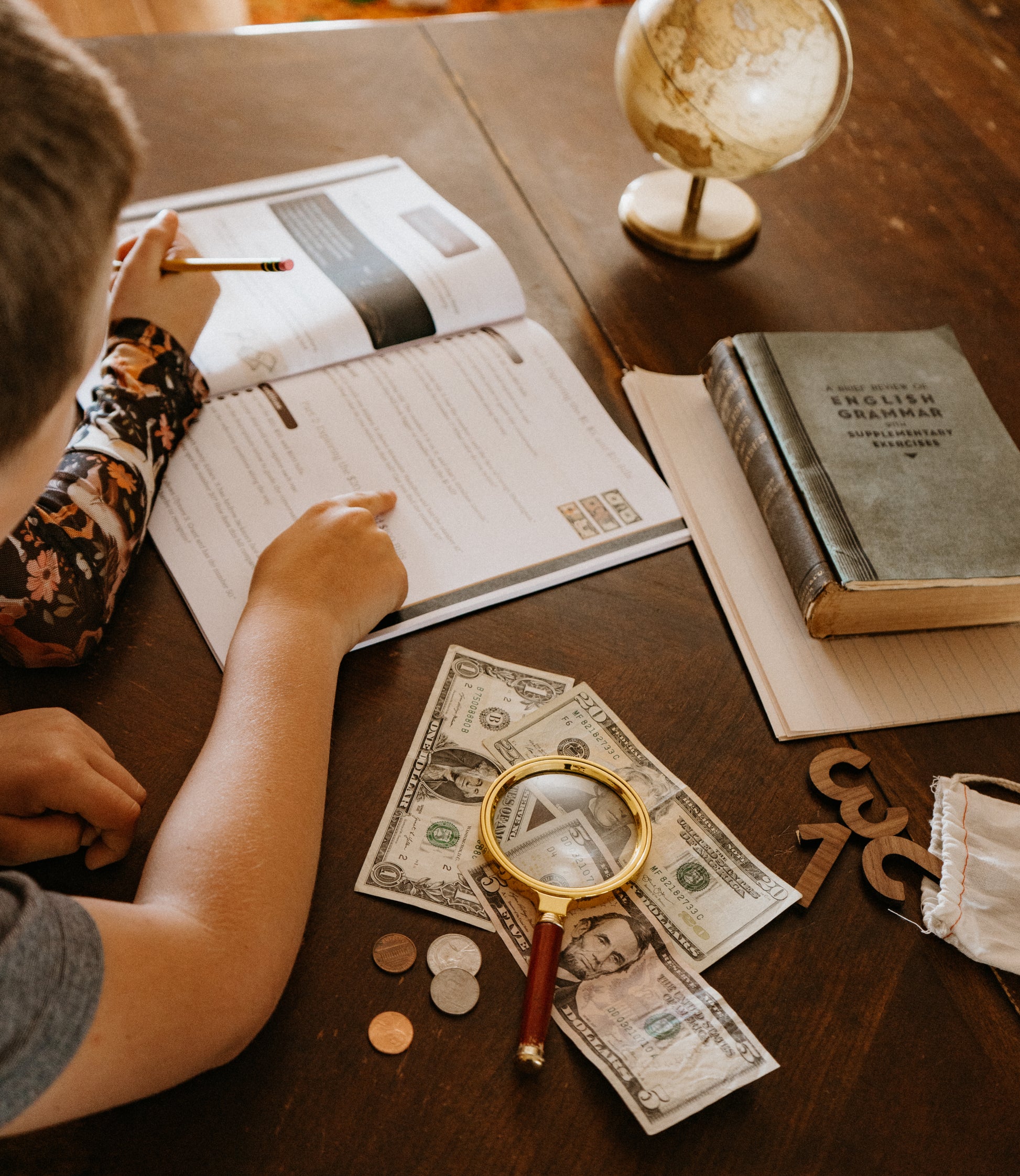 Children studying with books, money, and a magnifying glass on a wooden table.